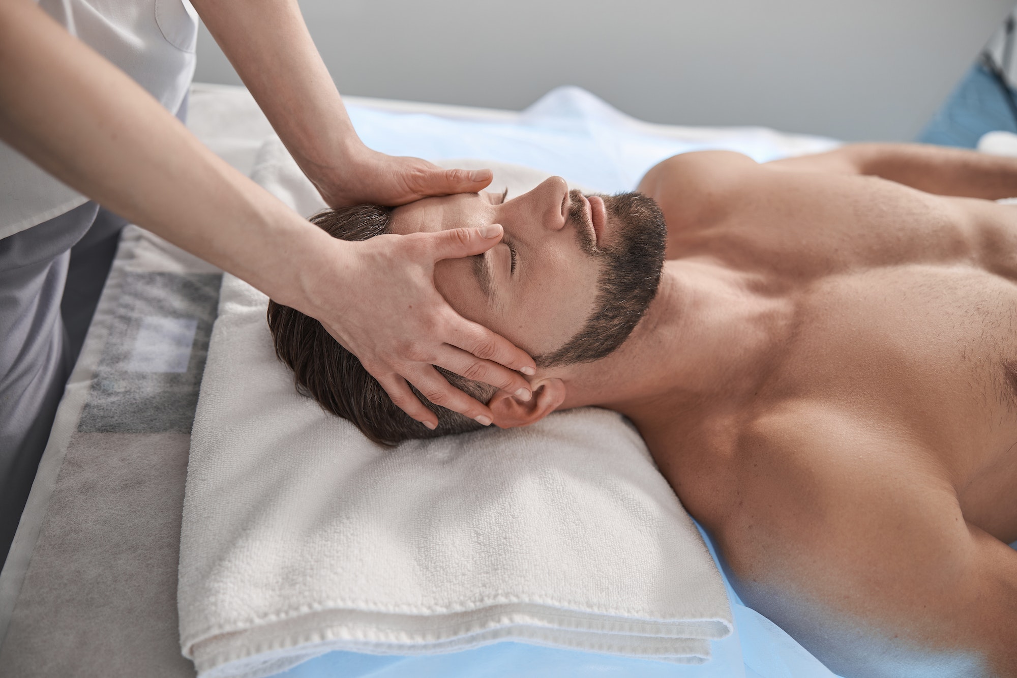 Massage therapist works with head of male lient lying on couch in contemporary clinic